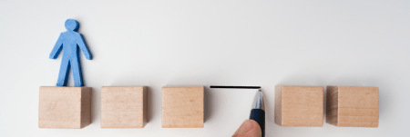 Five wooden blocks are lined up with a gap between them. A blue paper cutout of a person stands on the first wooden block.