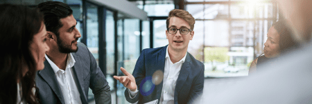 A group of professionals sit in a meeting. One person leads the conversation and the others listen.