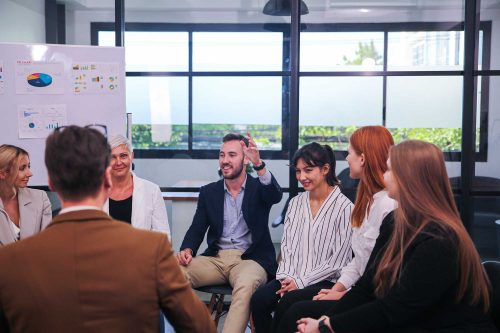 Development Solutions Team members sitting in a circle, actively discussing ideas near a whiteboard with charts in a bright office.