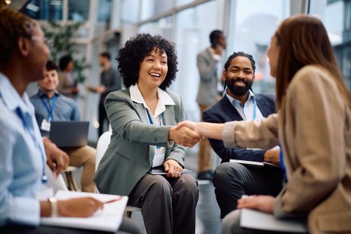 Selection Solutions Diverse professionals at a networking event, smiling as two women shake hands in a modern office setting.