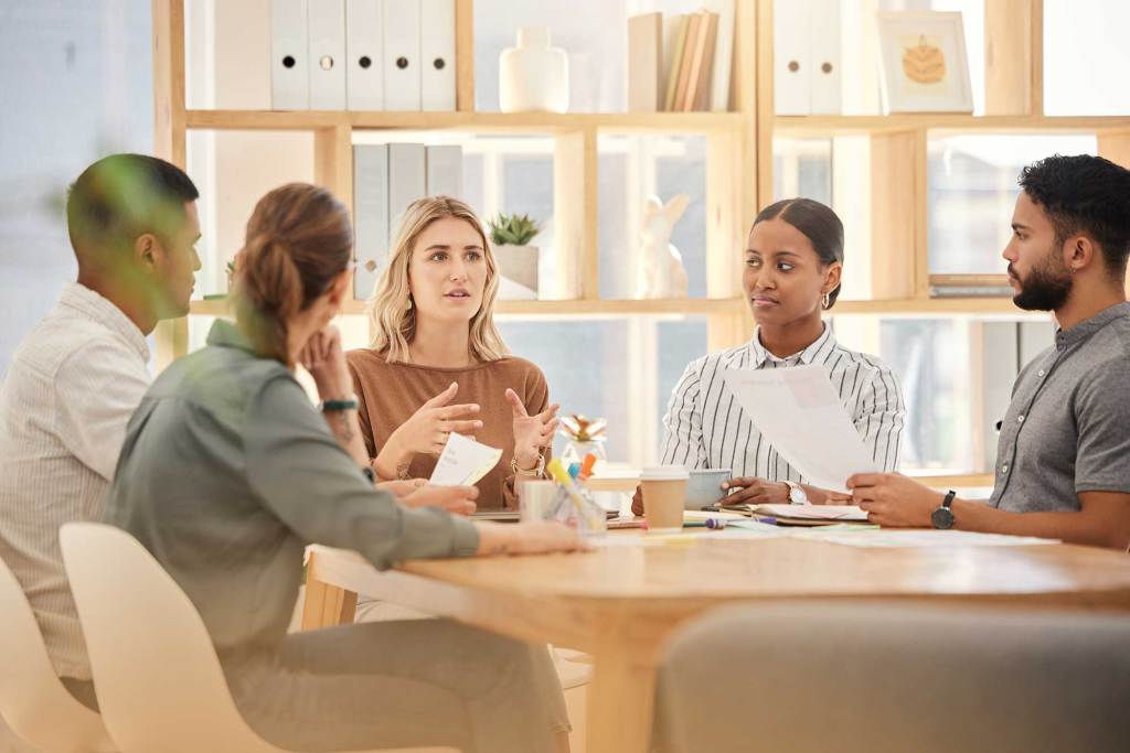 Succession Planning Solutions Five people having a meeting around a wooden table in an office, with documents in hand and shelves in the background.