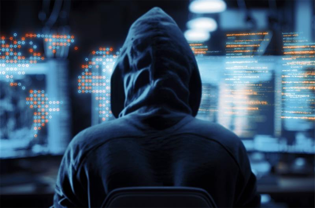A person in a hooded sweatshirt sits in a dark room facing multiple computer screens displaying code and digital graphics, suggesting a cybersecurity environment.
