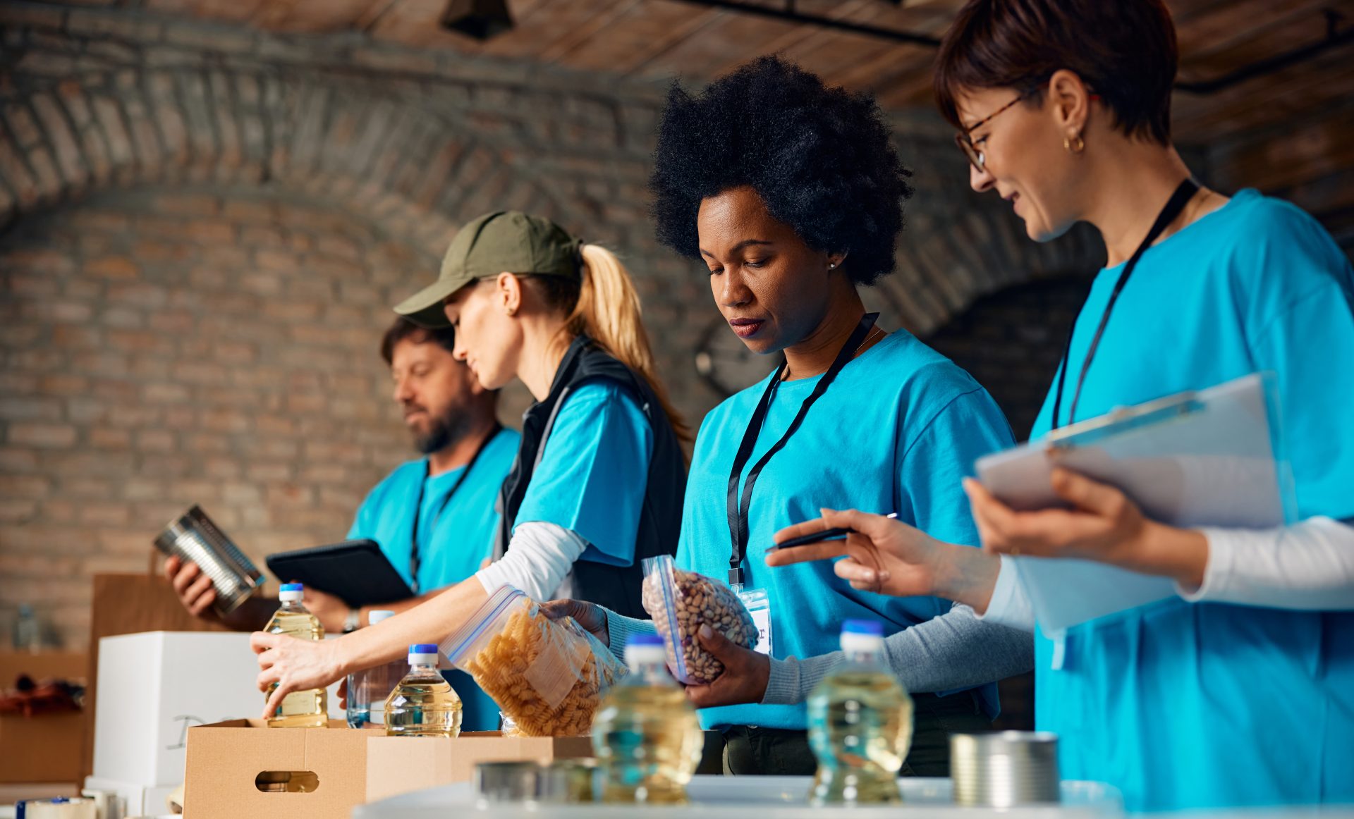Volunteers wearing matching blue shirts sort and pack food items while checking inventory on clipboards and a tablet.